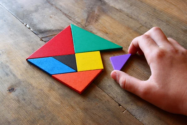 Man's hand holding a missing piece in a square tangram puzzle, over wooden table. — Stock Photo, Image