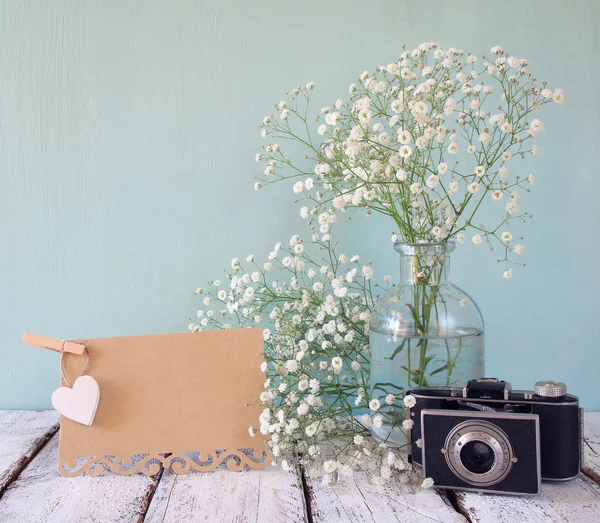 Fresh white flowers, heart next to vintage empty card and old camera over wooden table. — Stok fotoğraf