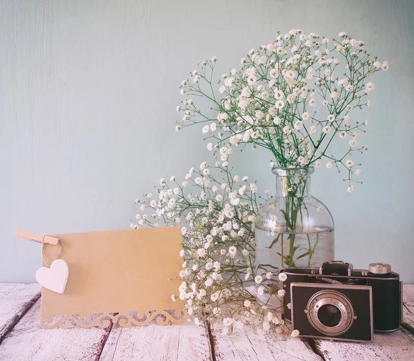 fresh white flowers, heart next to vintage empty card and old camera over wooden table.