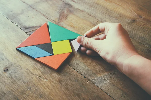 Mâna omului care ține o piesă lipsă într-un puzzle pătrat de tangram, peste masa de lemn . — Fotografie, imagine de stoc