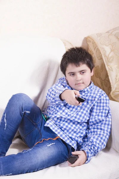 Un garçon en jean et chemise assis sur le canapé et appuie sur le bouton de la télécommande — Photo