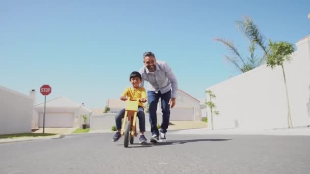 快乐的拉丁父亲帮助微笑的男孩在街上骑木制的平衡自行车 快乐的中东孩子和年轻的爸爸骑自行车在车道上 笑爸爸教儿子骑平衡自行车 — 图库视频影像