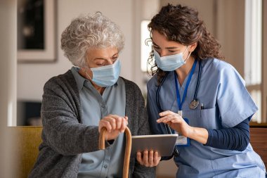 Genç doktor ve yaşlı kadın, ev ziyareti sırasında yüz koruyucu maske takarak dijital tablet üzerinde tıbbi kayıtları inceliyorlar. Elinde baston tutan yaşlı kadın, hemşire ile oturup kovid-19 terapisini dinliyor. Ameliyat giyen olgun bir kadın.