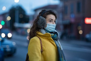 Gün batımında dışarıda tıbbi koruma maskesi takan bir kadın. Kirliliğe karşı maske takan genç bir kadın kış akşamı dışarıda dikiliyor. Coronavirus salgını sırasında, Winter City caddesinde gri kulaklı bir kız..