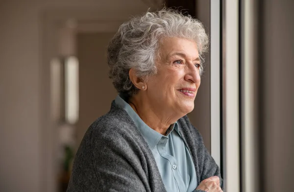 窓から見える白髪の老女の笑顔 Covid 19による隔離中に窓の近くに立つ幸せな先輩女性 外を見ながらセーターを着て引退し 瞑想的な女性 — ストック写真