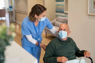 Koruyucu yüz maskesi takan dost canlısı hemşire, kovid19 salgını sırasında tekerlekli sandalyedeki yaşlı adama yardım ediyor. Cerrahi maske takan genç hemşire Coronavirus salgını sırasında tekerlekli sandalyede oturan yaşlı bir hastaya bakıyor.