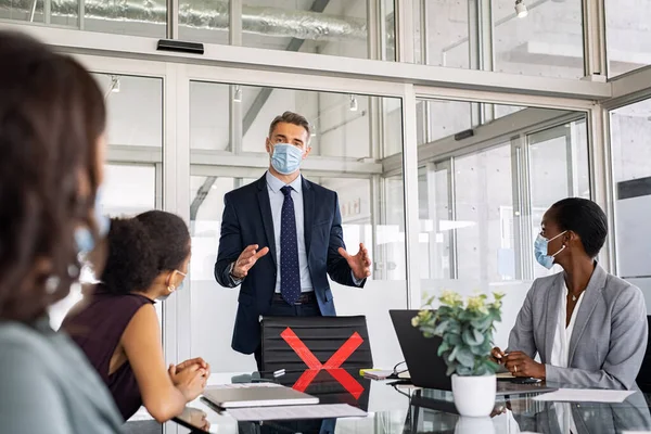 Mature businessman discussing project details with colleagues in modern office with face mask for safety against covid-19 virus. Business people brainstorming and discussing in conference room wearing surgical face mask for covid19 pandemic. Seriuos
