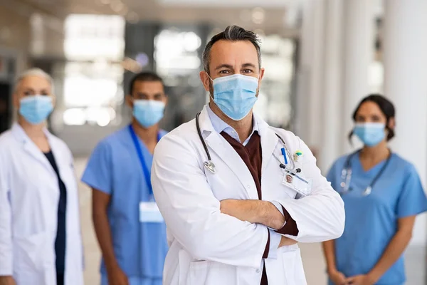 Portrait of happy mature doctor standing in corridor with medical team at hospital wearing surgical face mask due to covid. Smiling general practitioner with crossed arms looking at camera and wearing labcoat with face mask for covid-19 pandemic. Por