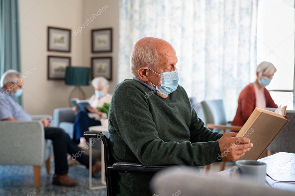 Senior man wearing protective mask and reading a book at nursing home with people in background. Lonely old disabled man sitting on wheelchair reading book during covid-19 pandemic. Elder wearing surgical face mask and reading during lockdown due to 