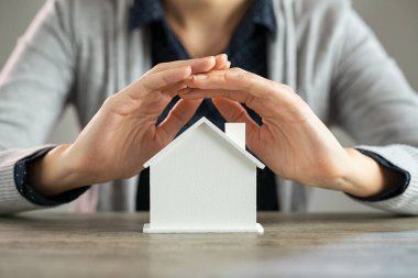 Masadaki beyaz ev modelini koruyan genç kadın ellerine yakın çekim. Masadaki karton figürün üzerinde el ele tutuşan kadının ayrıntıları. Kadın emlak yatırımını evinin ölçekli modeline göre koruyor..