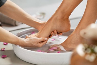 Genç bir kadının güzellik salonunda ayaklarının ovalanması. Genç masörün elleri kaplıcada ayaklarını suyla ve pembe taçyaprağıyla gri bir kapta yıkıyor. Lüks güzellik salonunda masaj yaptıran kız: ayak refleksolojisi ve ayak masajı.