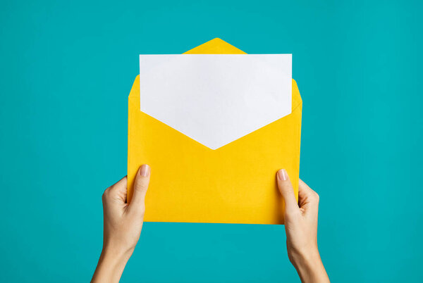 Women hand holding yellow envelope and card. Woman hands holding empty white card and open yellow envelope letter on blue background. Close up of girl showing empty letter ready for your text.