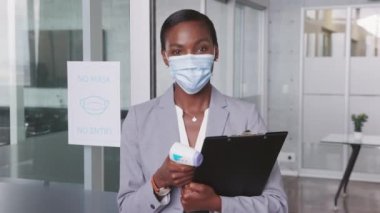 Ofise girmeden önce vücut ısısını kontrol etmek için kızılötesi alın termometresi kullanmaya hazır Afrikalı bir iş kadını. Salgın virüs salgını. Termometre tutan cerrahi maske takan siyahi iş kadınının portresi..