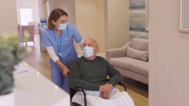 Koruyucu yüz maskesi takan dost canlısı hemşire, kovid19 salgını sırasında tekerlekli sandalyedeki yaşlı adama yardım ediyor. Cerrahi maske takan hemşire Coronavirus sırasında tekerlekli sandalyede oturan yaşlı bir hastaya bakıyor..