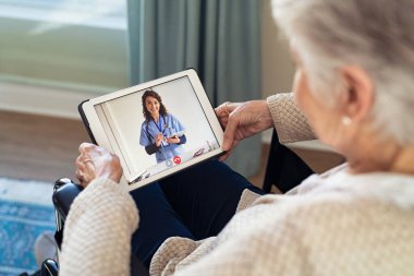 Tekerlekli sandalyede oturan yaşlı bir kadının, salgın sırasında evde otururken doktoruyla görüntülü konuşma yapması. Dijital tabletteki yaşlı kadınla video konferansında yardımcı olan pratisyen hekimi kapatın. İnternette hasta bir kadın var.