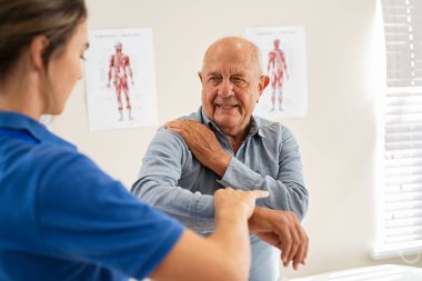 Genç kadın fizyoterapist yaşlı adama klinikte dirsek egzersizinde yardım ediyor. Genç bayan doktor yaşlı bir hastanın dirseğini kontrol ediyor. Yaşlı adam, özel klinikte profesyonel osteopatlarla görüşürken omuz ağrısına masaj yapıyor..