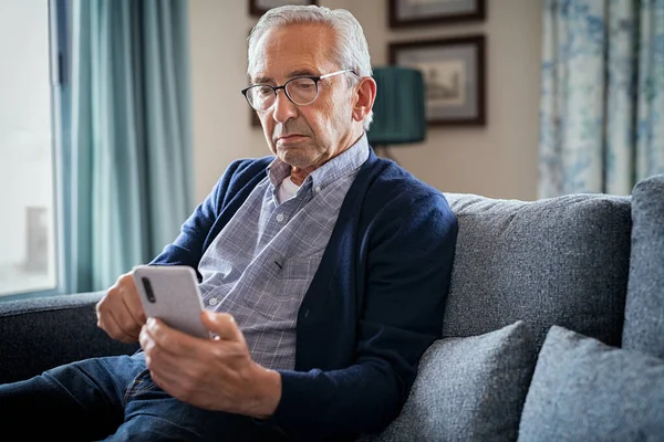老头儿戴着眼镜 在家里用智能手机 弄糊涂了 一个成熟的男人坐在沙发上很困难地在智能手机上读信息的画像 老年人很生气 一个人坐在客厅里 学习如何使用手机 — 图库照片