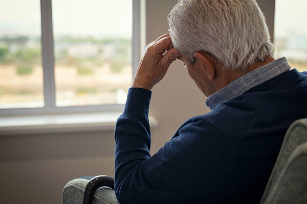Rear view of sad senior man sitting near window at home. Tired and depressed old man sitting on armchair in living room feeling hurt and lonely. Frustrated senior sitting on sofa at nursing home with head leaning on his hand while looking through the