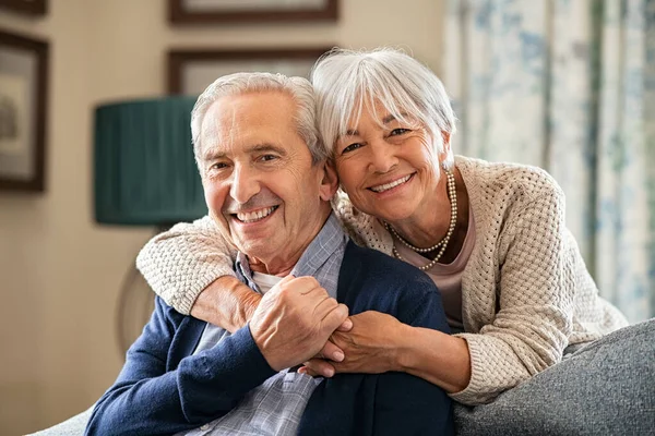 一个浪漫的老年男子和他美丽的妻子呆在家里的画像 坐在沙发上 从退休丈夫身后拥抱着一个笑容可亲的老妇人 快乐的老夫妇高兴地看着相机 — 图库照片