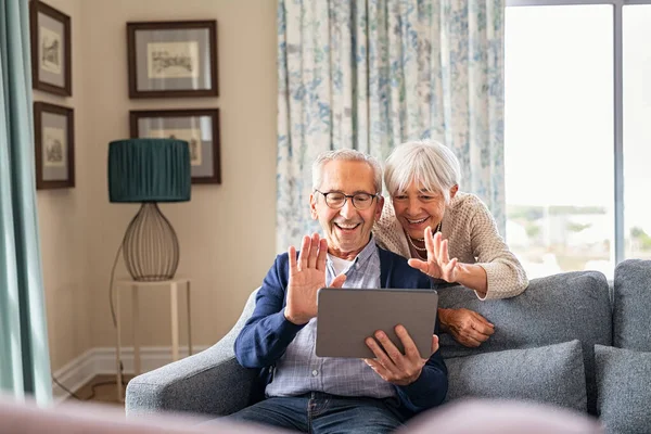 快乐的老夫妇使用数字平板电脑举行视频会议 一边在家里笑一边挥手 面带微笑的祖父母打电话问候坐在沙发上的家人 老人和快乐的女人用数码平板电脑和 — 图库照片