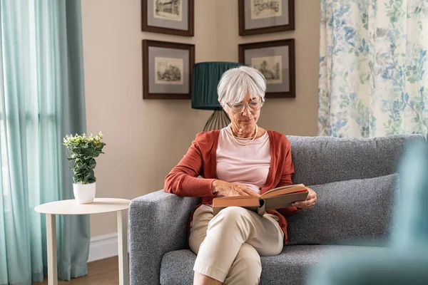 老太婆坐在沙发上看书 老太太一边在家里休息一边在沙发上看书 老奶奶一个人带着眼镜看小说 — 图库照片