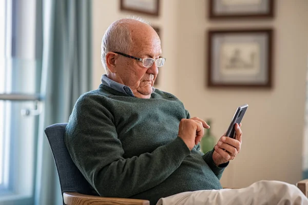 Old man wearing eyeglasses to use smartphone at home. Handsome senior man at home relaxing on couch reading a message on cellphone. Senior sitting alone in his living room and using mobile phone.