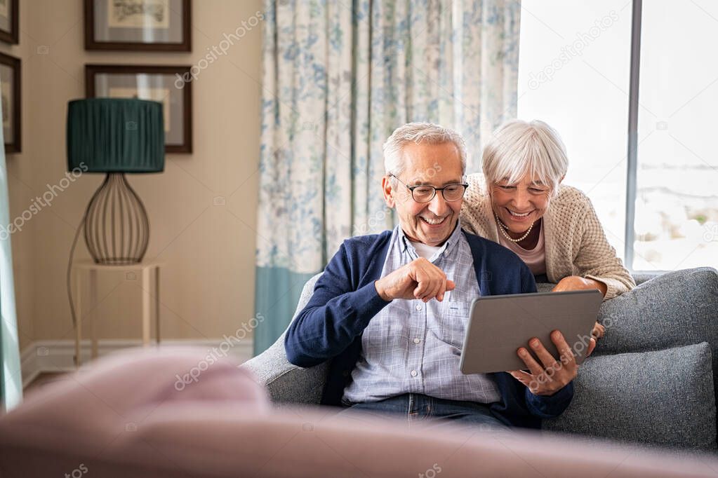 Happy old couple sitting on sofa using digital tablet with copy space. Cheerful senior man and beautiful elderly woman relaxing at home while watching funny video on digital tablet. Smiling grandparents making a video call with their nephews during l