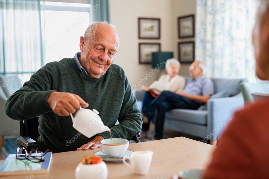 Smiling old man pouring hot tea in cup from kettle in nursing home. Elderly man in care centre sitting on wheelchair and drinking tea. Happy disabled elder in wheelchair enjoying tea time in the afternoon together with other patient of the care facil