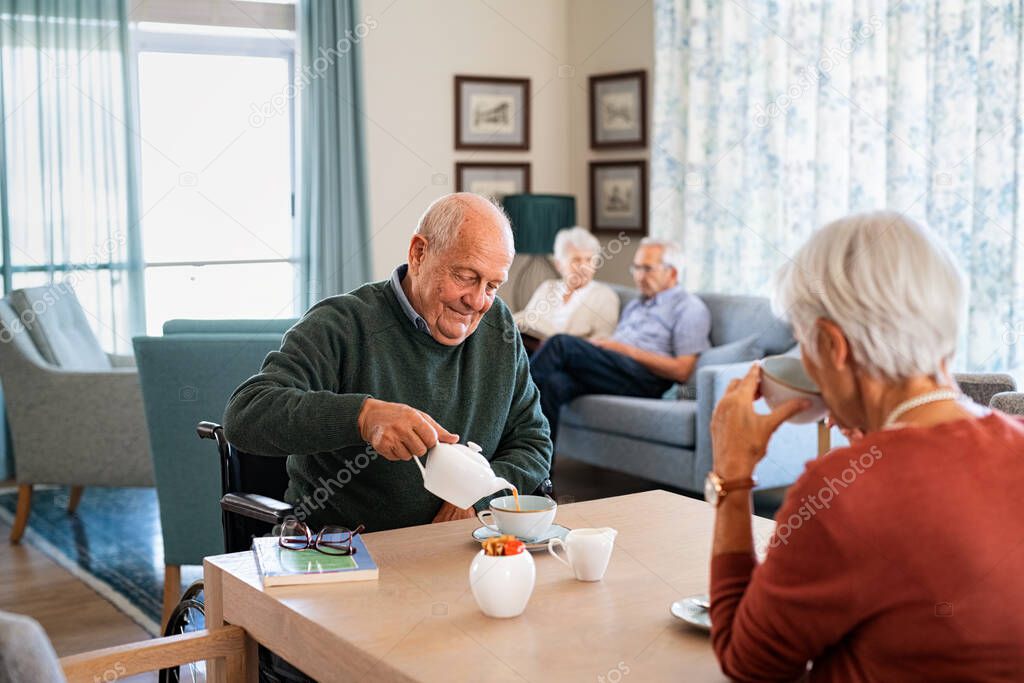 Smiling senior man pouring tea in cup from teapot in care facility with wife sitting at table in the common area. Elderly man in care centre sitting on wheelchair and serving tea. Serene disabled senior man enjoying tea time with friends during a vis