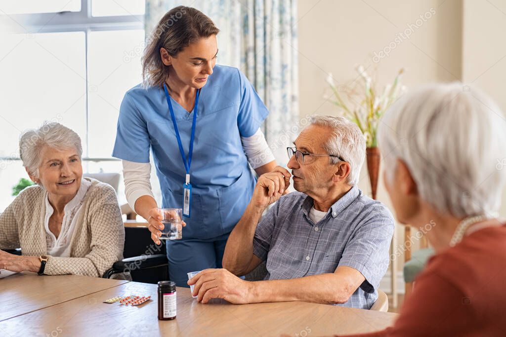 Young nurse in uniform giving medicine to group of seniors at modern nursing home. Caregiver giving medicine to old patient sitting at table at care facility centre. Elderly man taking pills from healthcare worker at retirement community together wit