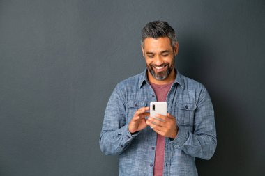 Gri arka planda izole edilmiş akıllı telefondan mesaj atan orta yaşlı, etnik çeşitlilikte bir adam. Gülümseyen Orta Doğulu adam gri duvara yaslanmış akıllı telefon kullanıyor. Mutlu melez adam cep telefonunda kopyalama alanı olan yeni bir uygulama kullanıyor..