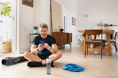 Evde egzersiz yaparken akıllı telefon kullanan olgun, yakışıklı bir adam. Yetişkin fitness adamı, evdeki akıllı telefondan günlük antrenmandan sonra kalp atışlarını kontrol ediyor. Formda, orta yaşlı bir adam cep telefonu kullanırken spor yaptıktan sonra dinleniyor..