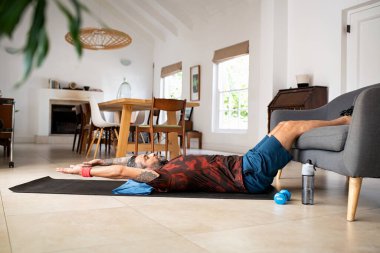 Olgun melez ırk adamı, evdeki kanepede bacaklarını tutarken yoga minderinde basın yapıyor. Orta yaşlı Hintli adam sabah rutininde mekik çalışıyor. Sağlıklı ve formda bir Orta Doğulu adam, kilitlenme sırasında evde karın kası egzersizi yapıyor.. 