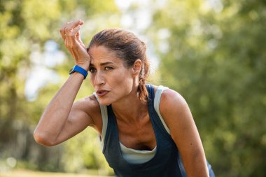Koşu yaptıktan sonra yorgun ve olgun bir kadın. Terli orta yaşlı atlet parkta fotokopi çekerek koşmaya ara veriyor. Yorgun orta yaşlı kadın yoğun fitness eğitiminden sonra ıslak alnını siliyor..