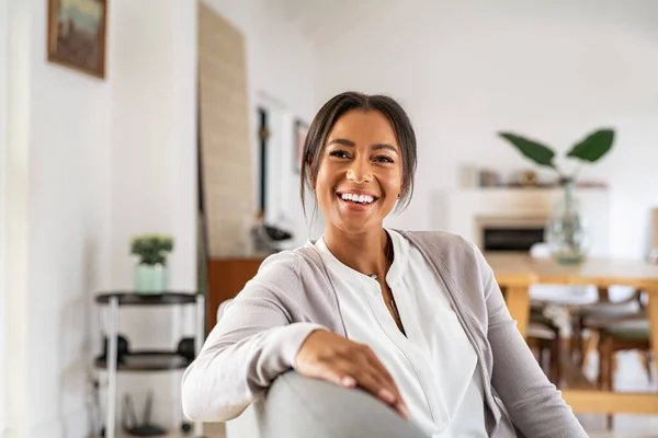 Happy mature woman relaxing on her couch at home in the living room. Beautiful young woman laughing alone in living room. Beautiful smiling african lady sitting on sofa.