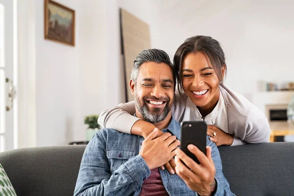 成熟的多种族夫妇在家里使用智能手机在一起和微笑 中年男子在沙发上放松 并在手机上向非洲裔美国妻子展示新的应用程序 中东男人和女人坐在家里的沙发上 用手机 — 图库照片