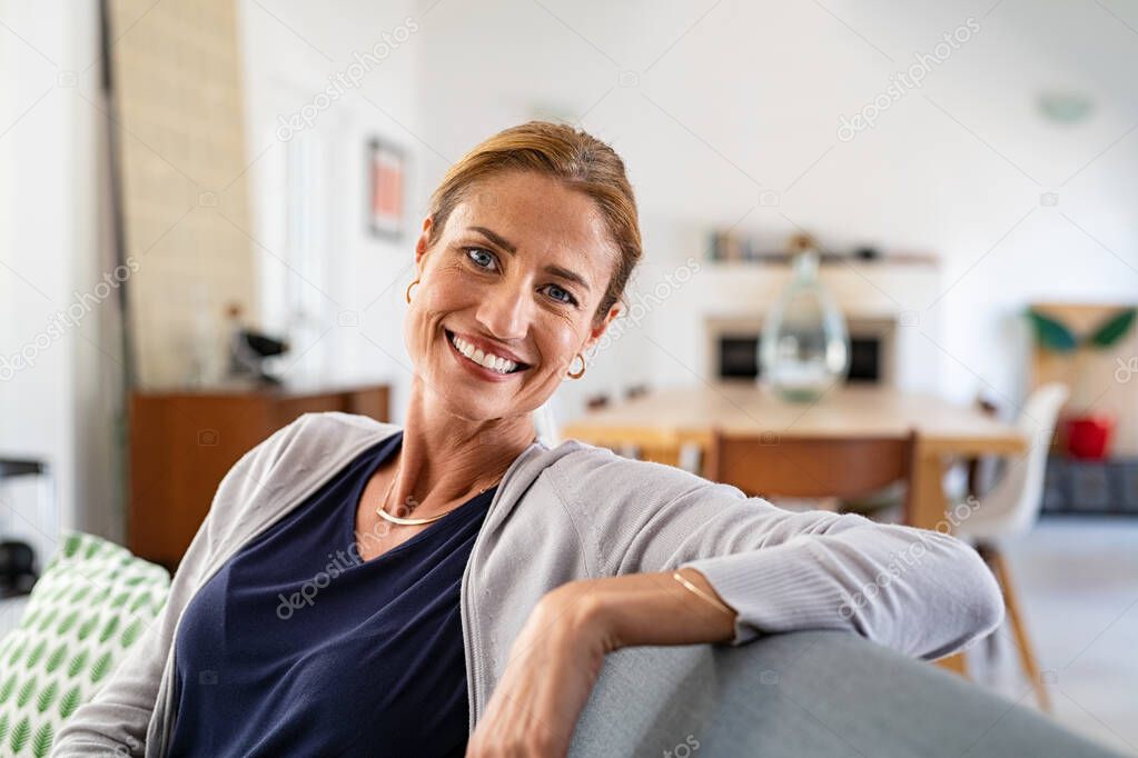 Happy mature woman relaxing on couch at home in living room. Close up face of middle aged woman looking at camera with copy space. Portrait of happy casual woman smiling and sitting on couch at home.