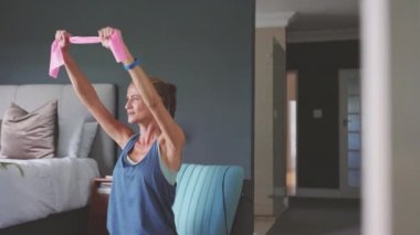 Orta yaşlı kararlı kadın pembe direniş bandı kullanarak koltukta oturuyor. Evde kilitli kalırken göğüs kasları için egzersiz yapan orta yaşlı fitness kadını. Yatak odasında direniş bandıyla çalışan sağlıklı, olgun bir bayan..