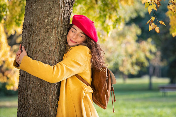 Beautiful girl with closed eyes hugging tree at park during autumn season. Beautiful woman hugging a tree and smiling outdoor. Casual cheerful woman embracing a tree trunk with a blissful expression on a sunny day: environmental conservation and natu