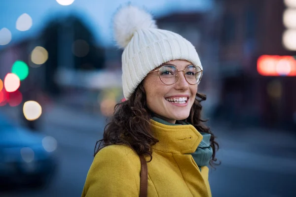 若い女性がバスを待っている街の通りに眼鏡をかけて暖かい帽子をかぶって笑顔 幸せな女の子は黄色のコートを着て バス停で冬の夜に屋外に立っている ケアフリー幸せな女の子で冷たい都市部で夕暮れ時 — ストック写真
