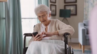 Tekerlekli sandalyede oturan gözlüklü yaşlı bir kadın akıllı telefon kullanıyor. Huzurevinde cep telefonuyla mesajlaşan kafası karışmış yaşlı bir kadın. Üst düzey hasta torunları ve ailesiyle iletişim kurmak için cep telefonu kullanıyor..