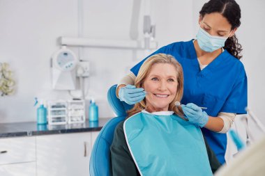 Modern bir klinikte dişçi muayenesi sırasında gülümseyen yaşlı bir kadın. Olgun hastaları muayene ederken dişçi aletleri kullanan çok ırklı genç bir dişçi. Ağız muayenesi ya da nüshalı diş hijyeni için sandalyede oturan yaşlı kadın..