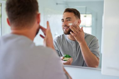 Yakışıklı bir adamın sabah rutinine bakarken yüzüne nemlendirici sürüşünün yansıması. Adam banyoda cilt bakımı sırasında yanağına nazikçe parmaklarıyla dokunuyor. Kendine güvenen, pijamalı, elinde krema kavanozu olan ve gülümseyen bir genç..