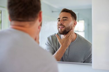 Genç bir adamın aynaya bakarken yüzüne dokunuşunun yansıması. Sakallı yakışıklı adam bakım sırasında muayene ediyor. Kendine güveni olan genç adam tıraş sonuçlarını kontrol ediyor..