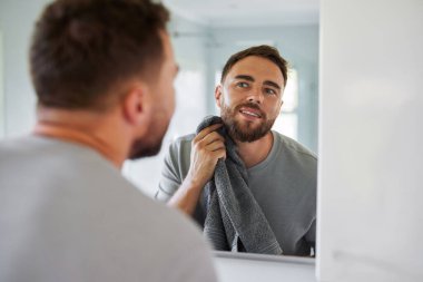 Genç bir adamın aynaya bakarken yüzünü havluyla kurulamasının yansıması. Yeni yıkandıktan sonra cildini okşayan sakallı yakışıklı bir adam. Banyoda cilt bakımı sırasında pijamalı, kendinden emin orta yaşlı bir adam..