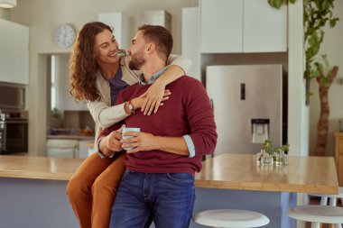 Mutlu çift, evde kucaklaşırken sabah çayının tadını çıkarıyor. Mutfak tezgahında oturan neşeli kadın kahve içerken kocasını kucaklıyor. Romantik bir çift birbirlerine günlük kıyafetlerle bakıyor..