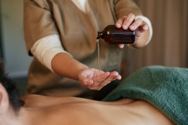 Profesyonel masözün elleri masaj için şişeden yağ döküyor. Masaj terapisti lüks tedavi seansı için eline cam şişeden yağ döküyor. Profesyonel masaj terapisti, gençleştirme bölümünde genç bir bayana masaj yapıyor.
