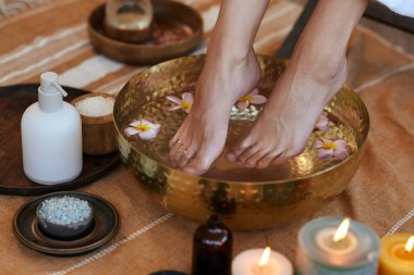 Pedikür seansı sırasında ayaklarında su ve frangipani çiçekleri olan altın bir kâse içinde genç bir kadının ayaklarını yakın çekim. Kadın, güzellik salonunda sıcak suyla lüks kasede dinleniyor. Kız, lüks bir kaplıcada ayak banyosuyla ısınıyor.