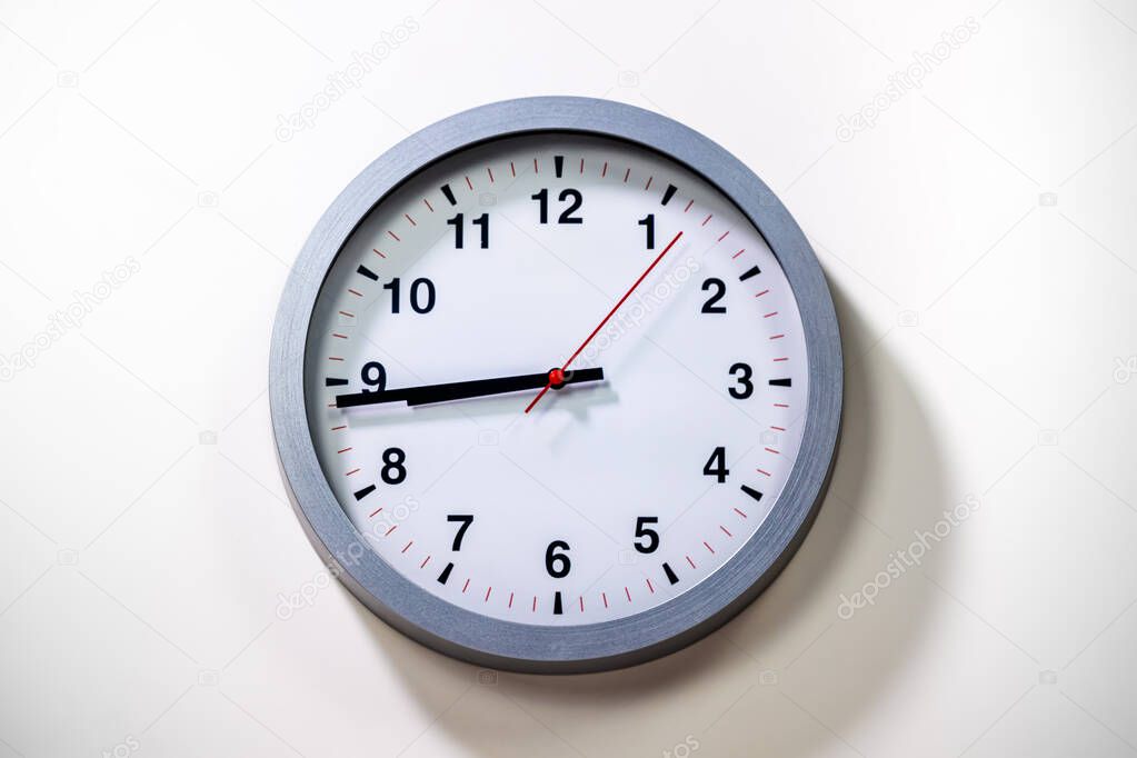 Grey Wall Clock show the time. Modern wall clock with grey frame on white background. Close up to a clock, with black time pointer and red second pointer. Shadow from the daylight.  