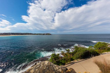 Bondi plajı etrafındaki kayalık kıyı şeridi, Sydney, Avustralya. Dalgalar sudaki kayalara çarpıyor. Tamarama, Tamarama sahili manzaralı. Mavi gökyüzünde beyaz bulutlar. Yeni Güney Galler 'de Sonbahar